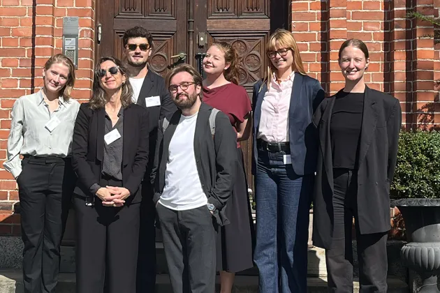 Seven people standing in the sun in front of a wooden door.