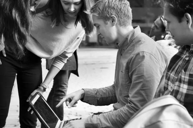Students gather round looking at a lap top. Photo.