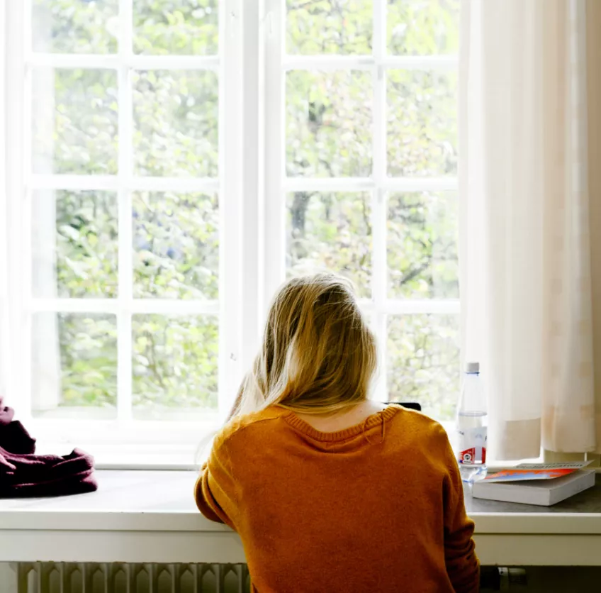 Student i orange tröja studerar vid fönster med ryggen mot kameran.